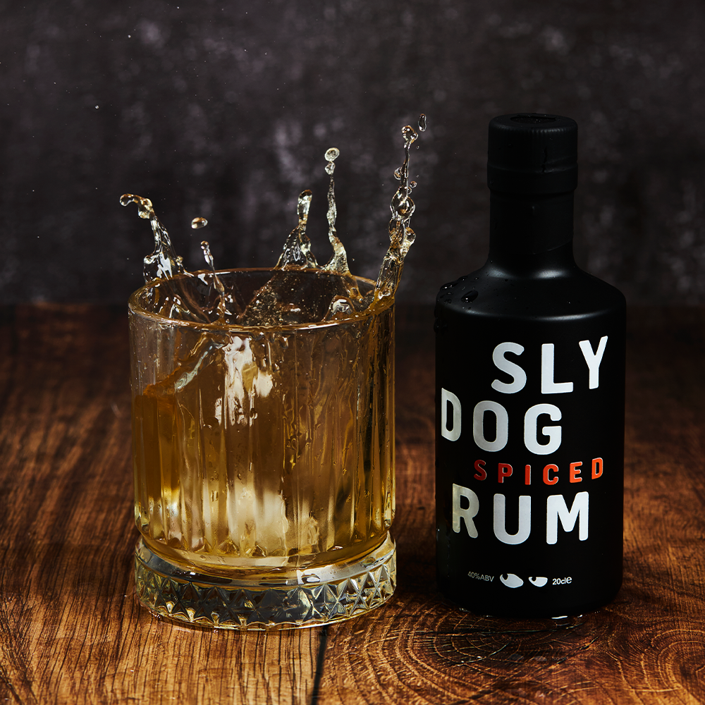 ice cube dropping into a small tumbler glass filled with rum, which a small 20cl bottle of SLY DOG Rum next to it 