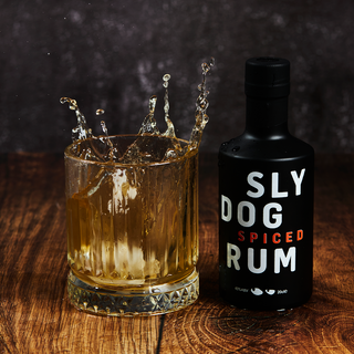 ice cube dropping into a small tumbler glass filled with rum, which a small 20cl bottle of SLY DOG Rum next to it 